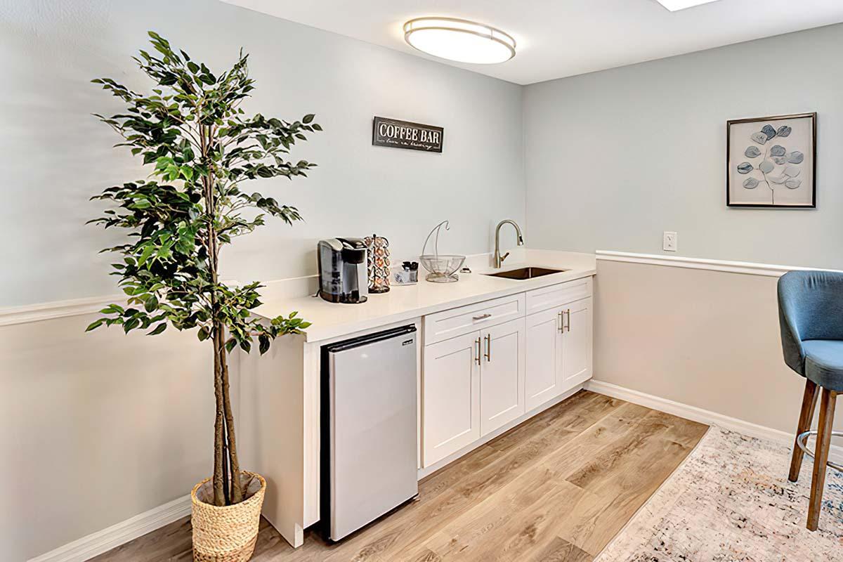A modern kitchenette featuring a small sink, a coffee machine, and a refrigerator. A decorative plant is in the corner, and a framed artwork hangs on the wall. The area is well-lit and has a clean, minimalist design with light-colored cabinetry and wooden flooring.