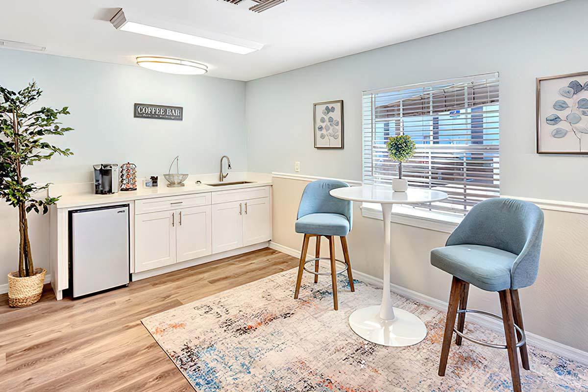 A bright, modern coffee bar area featuring a small round table with two blue upholstered stools, a white cabinet with a sink, a coffee machine, and a refrigerator. The walls are painted light blue, and there are decorative prints and a potted plant in the corner, with a colorful area rug on the floor.