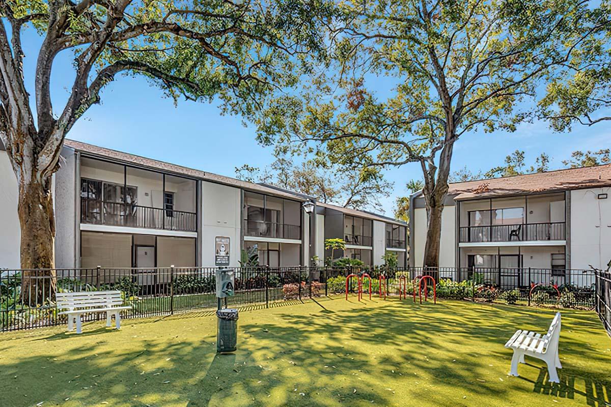 A landscaped courtyard of an apartment complex featuring green grass, benches, and exercise equipment surrounded by trees. The buildings are two stories high with balconies, and the area is enclosed by a fence, creating a welcoming outdoor space for residents to enjoy.