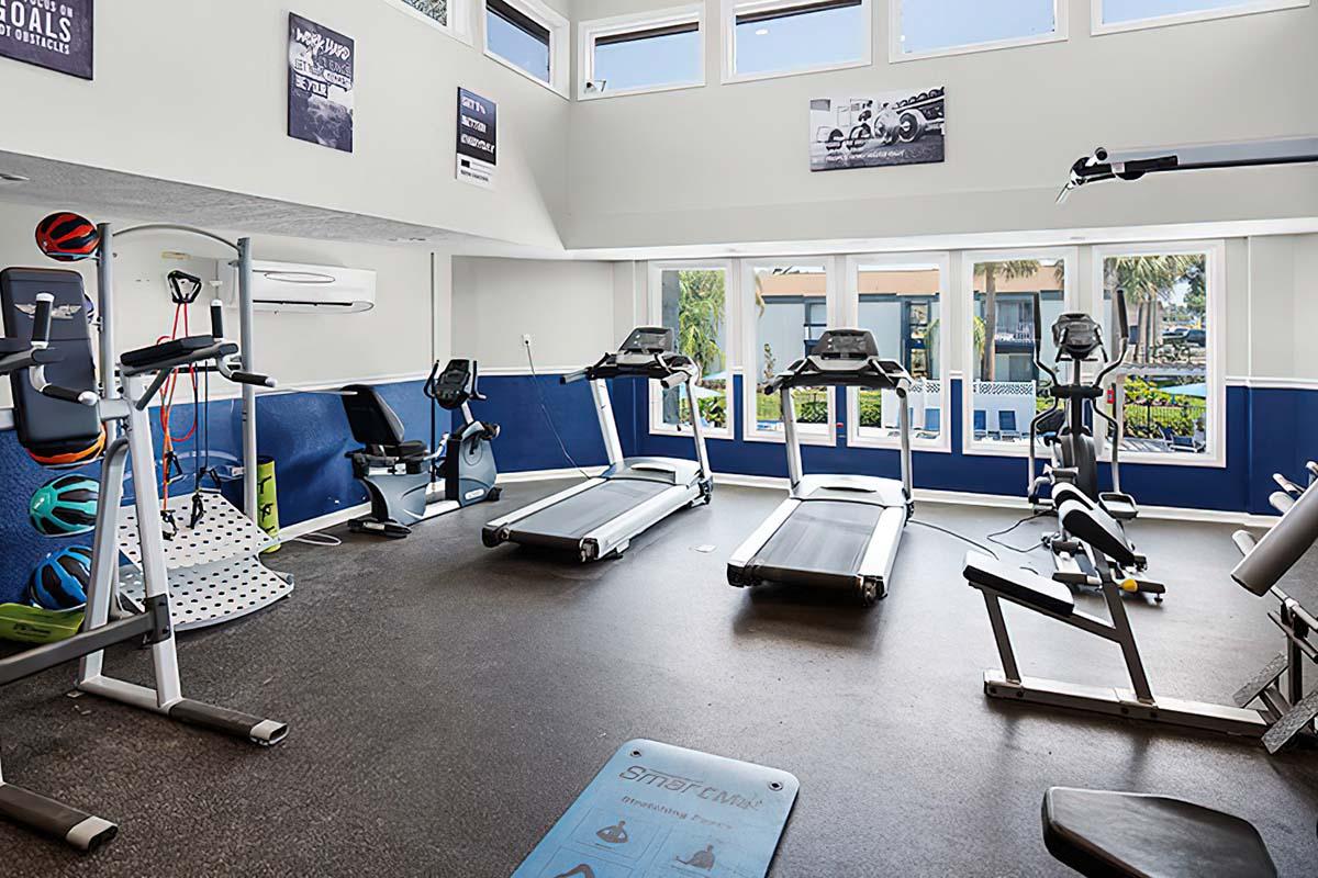 A bright and modern gym space featuring multiple treadmills, a stationary bike, and strength training equipment. Large windows provide natural light, and motivational posters decorate the walls. The floor is clean and there are exercise mats nearby, creating an inviting atmosphere for workouts.