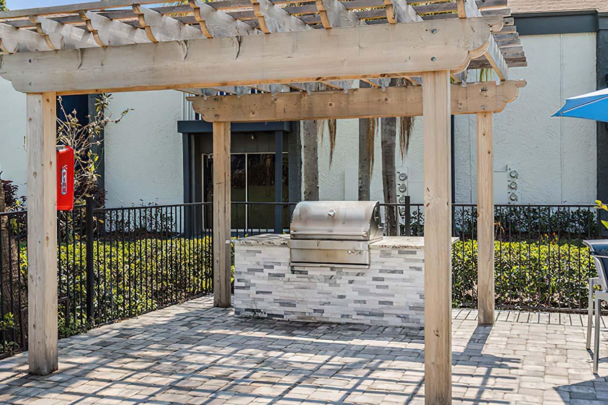 An outdoor grilling area featuring a stainless steel grill mounted on a stone countertop, sheltered by a wooden pergola. Surrounding the area are landscaped plants and a black fence, with seating visible in the background under a blue umbrella.