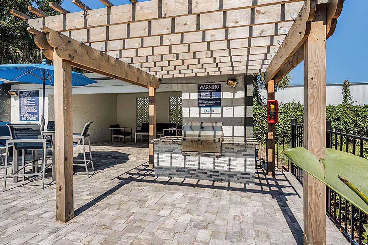 A shaded outdoor area with a wooden pergola, featuring seating arrangements with gray chairs and umbrellas. A stone countertop has an outdoor grill or barbecue setup, and safety signage is visible on a wall. The background includes greenery and blue sky.