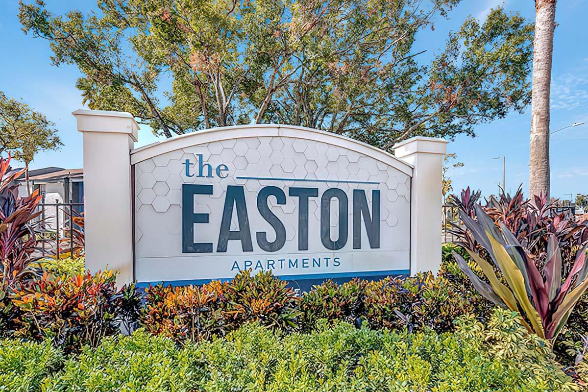 Sign for The Easton Apartments, displaying the name prominently against a decorative backdrop of colorful landscaping and trees under a clear blue sky.