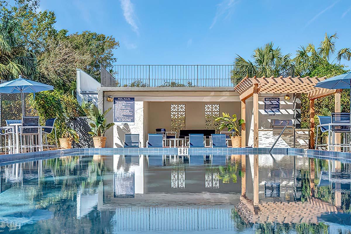 A clear blue swimming pool reflects the surrounding greenery and a sunny sky. In the background, there is a sheltered seating area with chairs and tables, along with a wall displaying informational signs. Palm trees and a sun umbrella enhance the tropical ambiance.