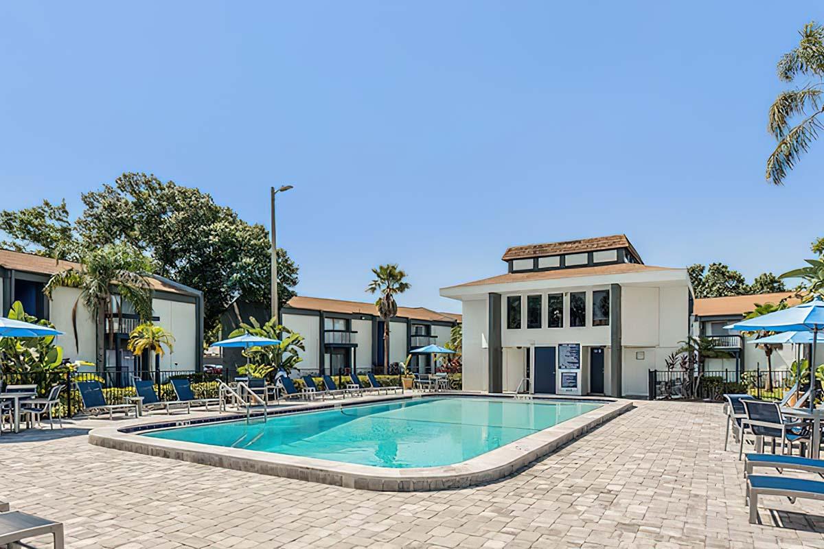 A clear blue sky over a swimming pool surrounded by lounge chairs and umbrellas. The pool area is bordered by palm trees and modern buildings in the background, featuring large windows and a welcoming entrance. The setting appears inviting and ideal for relaxation.