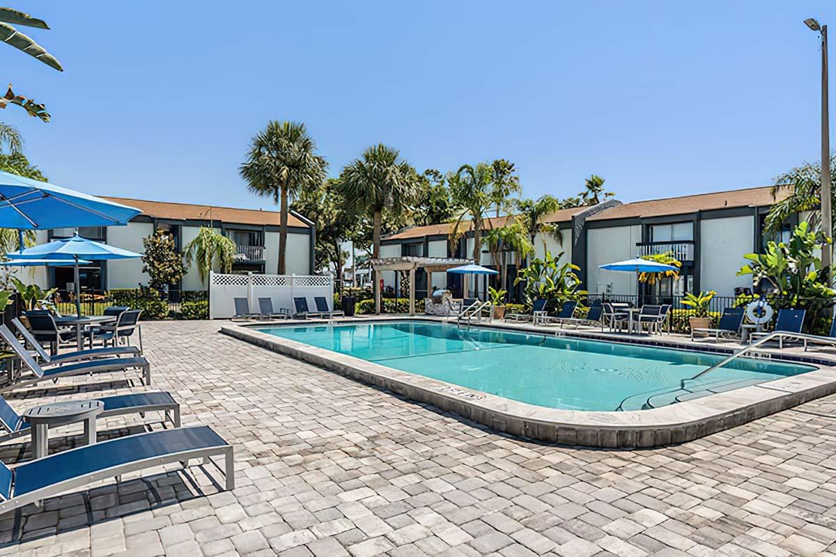 A sunny outdoor pool area surrounded by palm trees and lounge chairs. Blue umbrellas provide shade for guests. The pool is clear and inviting, with a few tables nearby. The backdrop features modern buildings and lush greenery, creating a relaxing atmosphere for leisure and enjoyment.