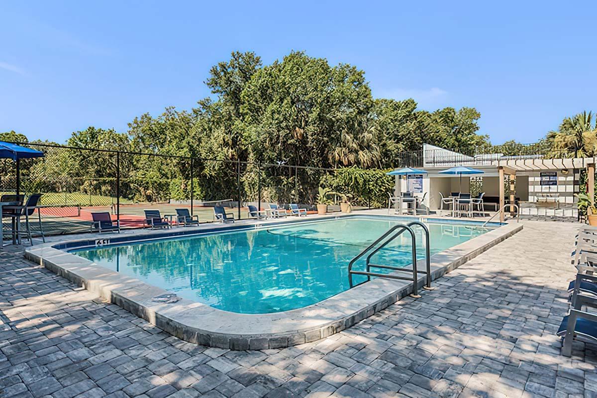A clear blue swimming pool surrounded by a paved area with lounge chairs. In the background, there are trees and a tennis court visible. To the side, there is a small building with outdoor seating, umbrellas, and a pergola for shade. The sky is clear and sunny, creating a relaxing atmosphere.
