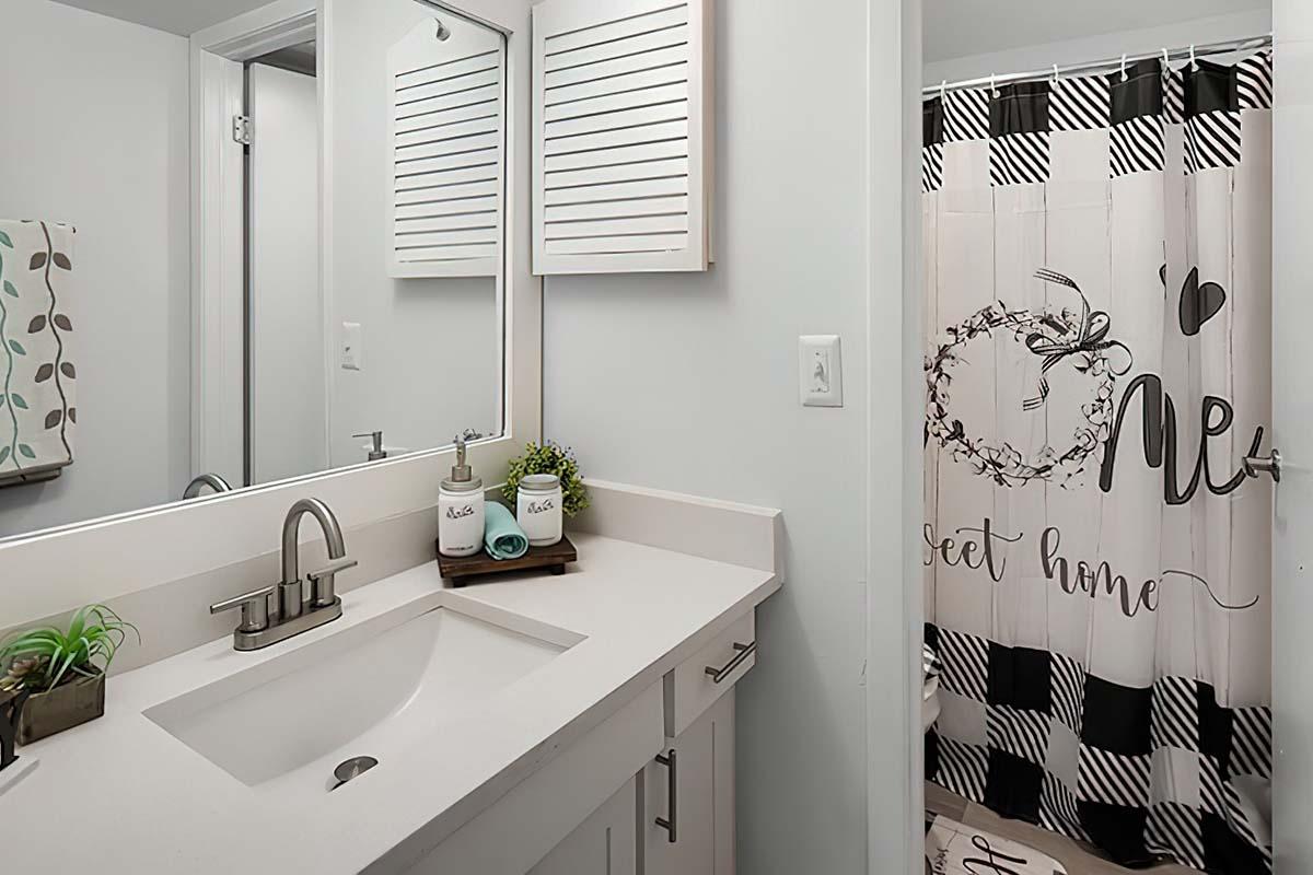 A clean, modern bathroom featuring a white vanity with a sink, a large mirror above, and a decorative plant. To the right, there's a shower with a black and white checkered curtain that reads "Home Sweet Home." The space has a bright and inviting ambiance.