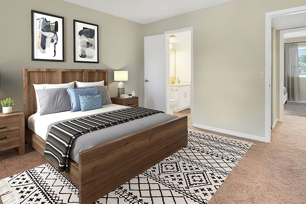 A cozy bedroom featuring a wooden bed with gray and white bedding, accented by a blue throw pillow. There are two framed artworks on the wall, nightstands with lamps, and a patterned area rug. A door leads to a bathroom, and a doorway connects to another room, showcasing a light and inviting atmosphere.