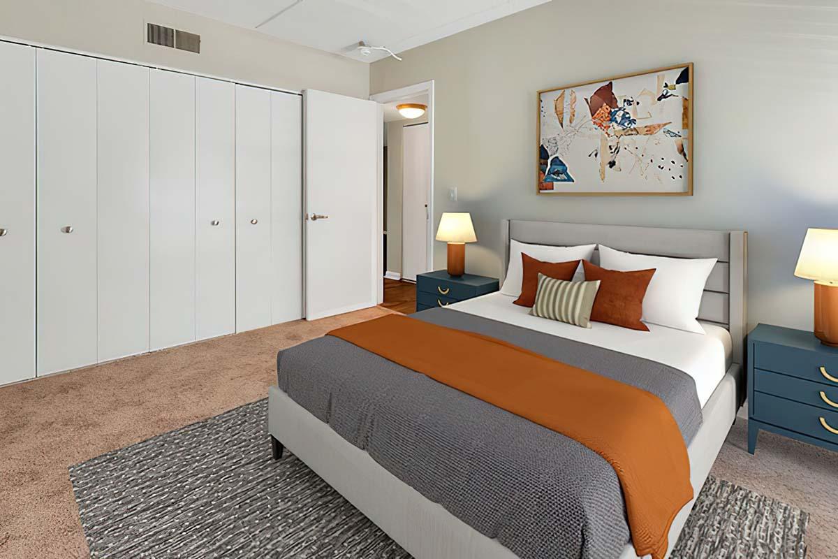 A bright bedroom featuring a queen-sized bed with orange and gray bedding, two bedside lamps, and blue nightstands. Walls are light-colored, and there is a large closet with white doors. A modern piece of art decorates one wall, and a textured area rug adds warmth to the carpeted floor.
