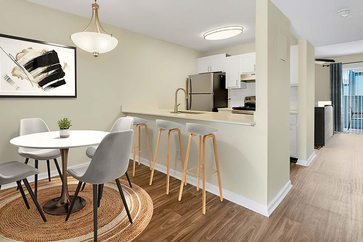 A modern open-concept kitchen and dining area featuring a round white table with four gray chairs, illuminated by a pendant light. There’s a decorative rug on a wooden floor, and a kitchen island with three high stools. The kitchen includes stainless steel appliances, and a large window offers natural light.