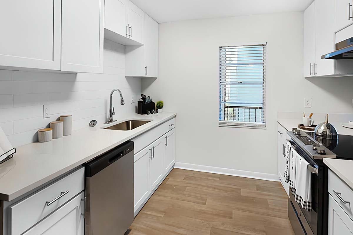 Modern kitchen featuring white cabinetry, a stainless steel sink, and an open window revealing a view. The countertop is light-colored with minimal decorative items. A stainless steel dishwasher and stove are visible, along with a wooden floor that adds warmth to the space.