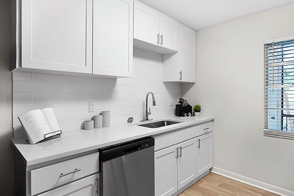 A modern kitchen featuring white cabinets, a stainless steel sink, and a dishwasher. There is a small book standing on the countertop, along with three decorative containers. A coffee maker is visible, and the area is well-lit with natural light coming through a window adorned with blinds.