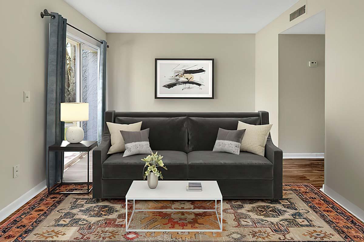 A modern living room featuring a dark gray sofa with decorative cushions, a coffee table with a vase of flowers, an area rug with intricate patterns, a lamp on a side table, and large windows with curtains. Wall art is displayed above the sofa, and wooden flooring is visible in the background.