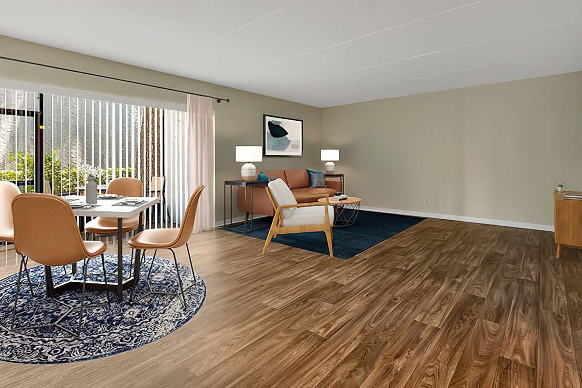 A modern living space featuring a dining table with four chairs, a cozy sofa, and two lamps. The room has large windows with light filtering through sheer curtains, a patterned area rug on hardwood flooring, and a stylish accent piece on a side table. Walls are painted light beige, giving a warm and inviting ambiance.