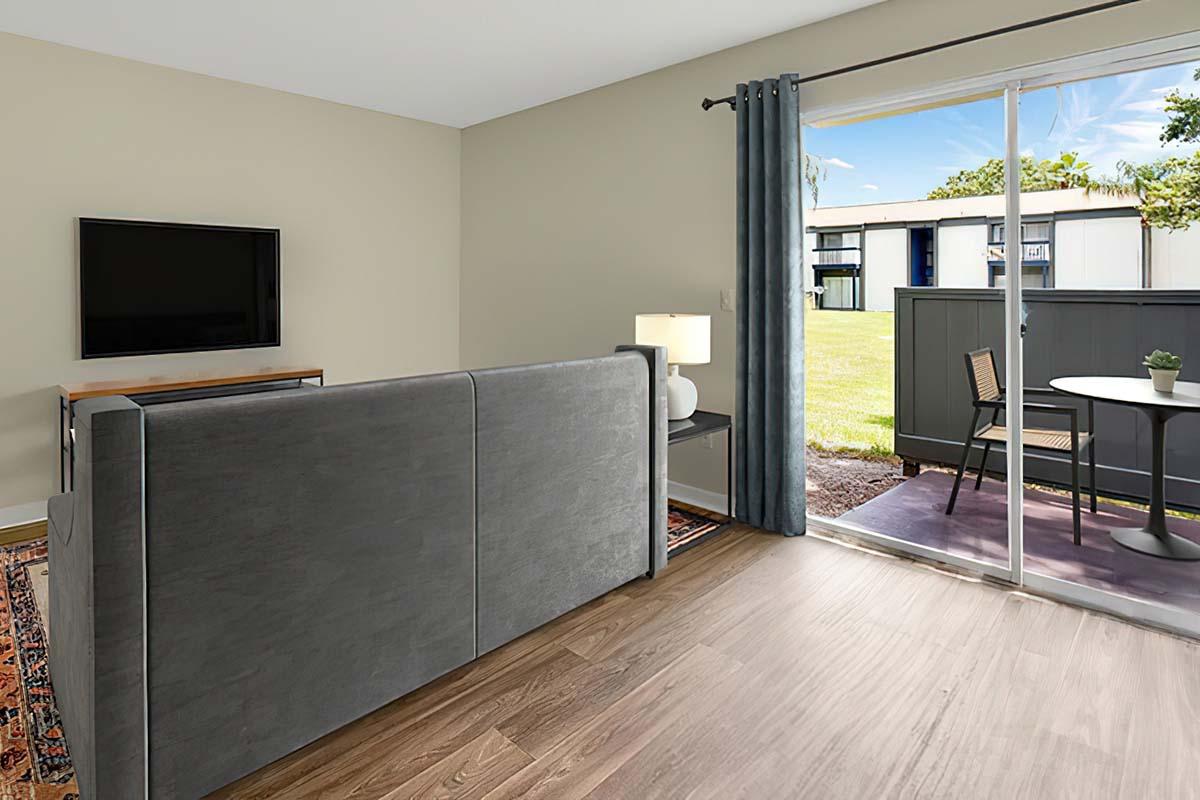 Interior view of a cozy living space featuring a gray couch, a small table with two chairs, and a flat-screen TV mounted on the wall. A sliding glass door leads to an outdoor area with grass and a small patio set. Natural light fills the room through the curtains. The floor has a warm wood finish.