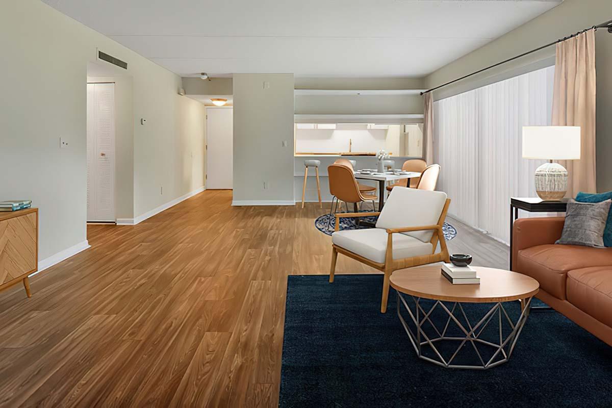 A spacious modern living room with hardwood floors, featuring a comfortable orange sofa, a white armchair, and a coffee table with a decorative bowl. There’s a dining area with a round table and chairs in the background, and large windows with sheer curtains allow natural light to fill the space.