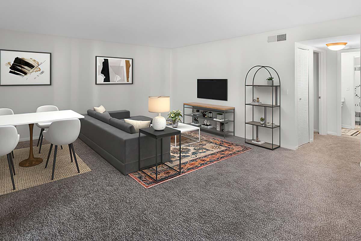 A modern living room and dining area featuring a gray sofa, a white dining table with chairs, a TV mounted on the wall, and a stylish shelf. The space has a neutral color palette, decorative art on the walls, a lamp on the side table, and a patterned rug on the carpeted floor, creating a cozy atmosphere.