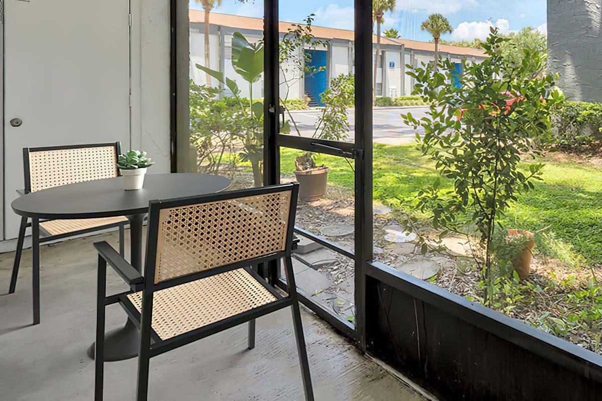 A view from a screened-in porch featuring a round black table and two woven chairs. Outside, there is a garden area with green grass, plants, and a glimpse of a parking lot and building in the background under a blue sky. A small potted plant is placed on the table.