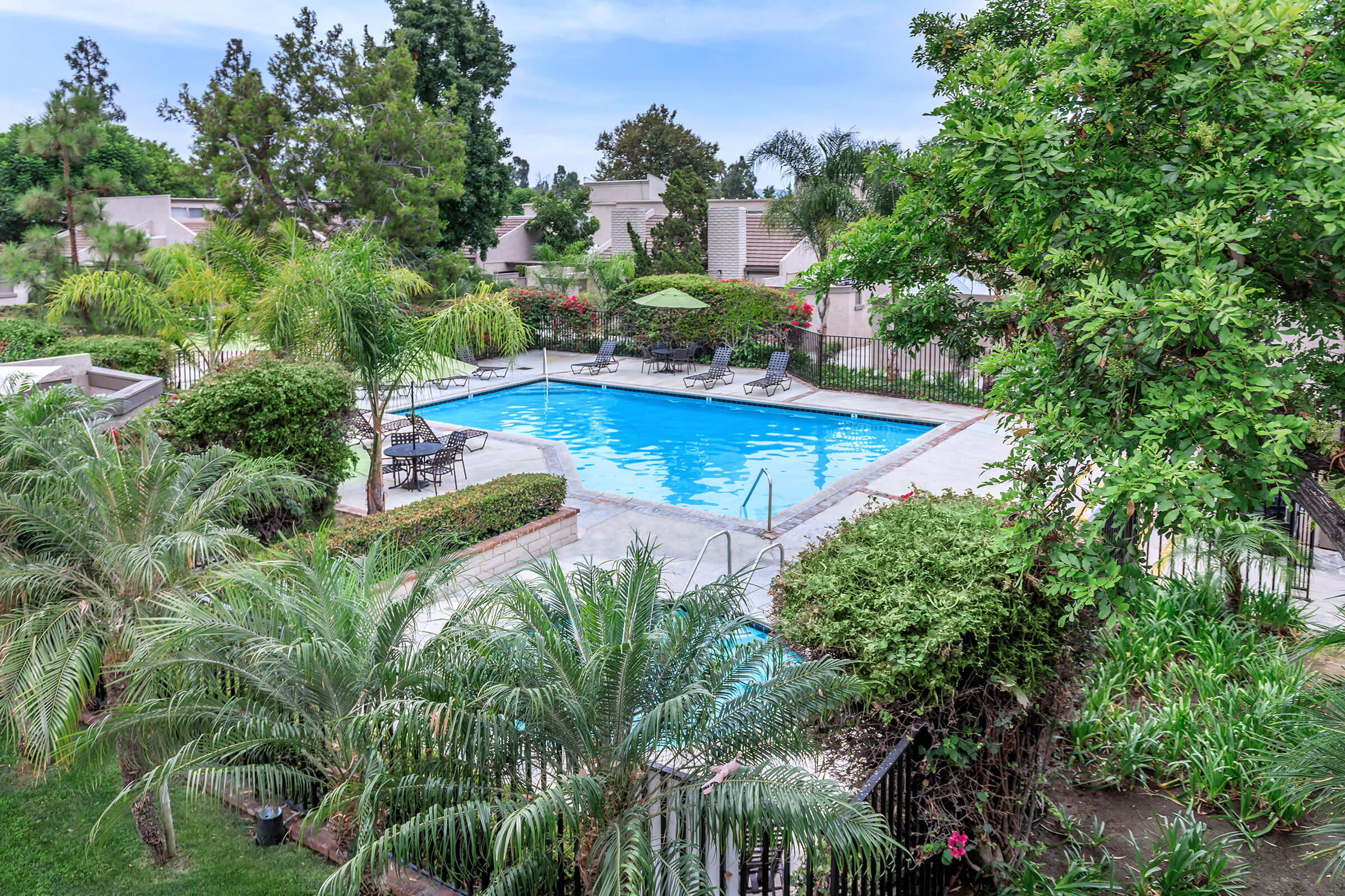 A serene outdoor pool surrounded by lush greenery, palm trees, and lounge chairs. The pool area features shaded umbrellas, with manicured hedges and shrubs in a tranquil residential setting.