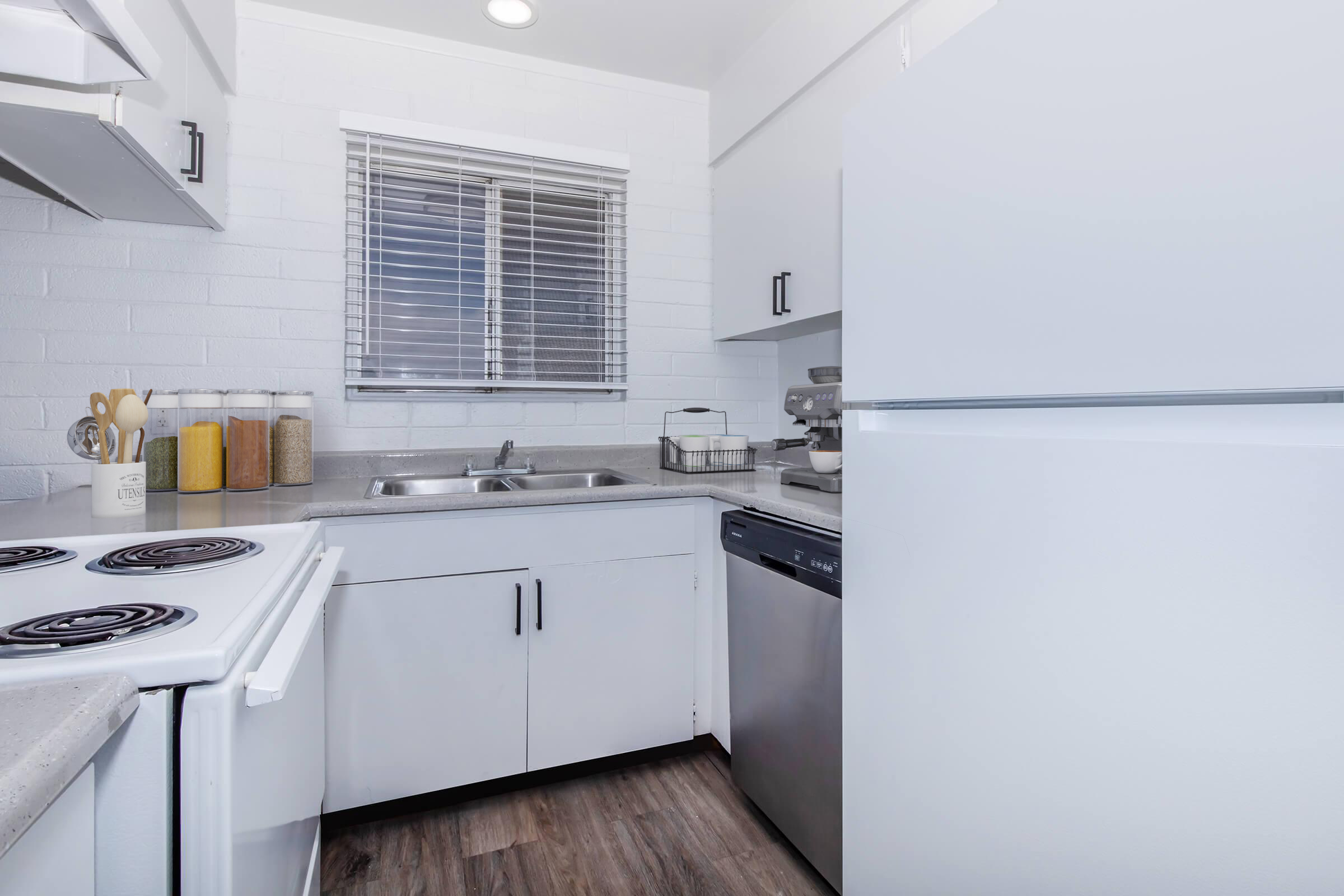 A modern kitchen featuring white cabinetry, stainless steel appliances, and a window with blinds. The space includes a sink, a stove with burners, a dishwasher, and organized kitchenware. The countertops are light-colored and there are jars for storage, creating a clean and functional atmosphere.