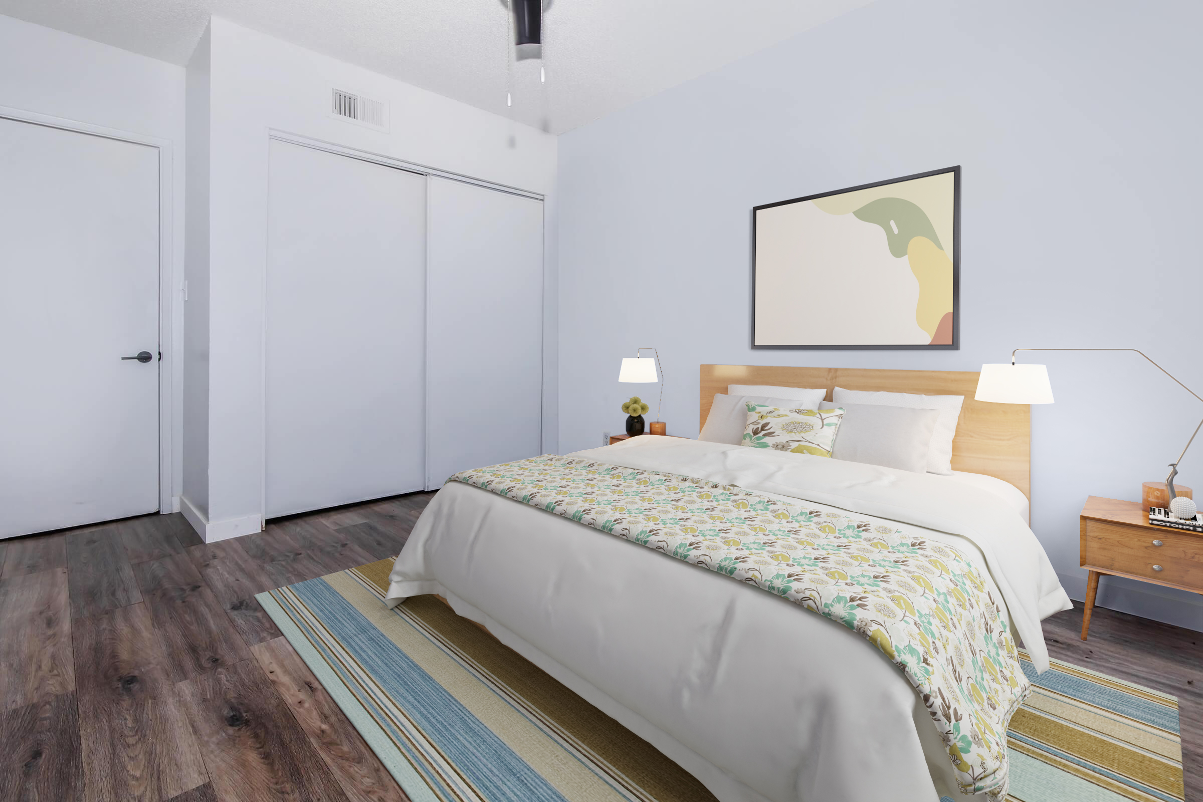 A cozy bedroom featuring a wooden bed with a colorful patterned blanket and decorative pillows. Two bedside lamps are on either side of the bed, and a modern artwork hangs above. The room has light blue walls, a striped area rug on the wooden floor, and a closet with sliding doors.