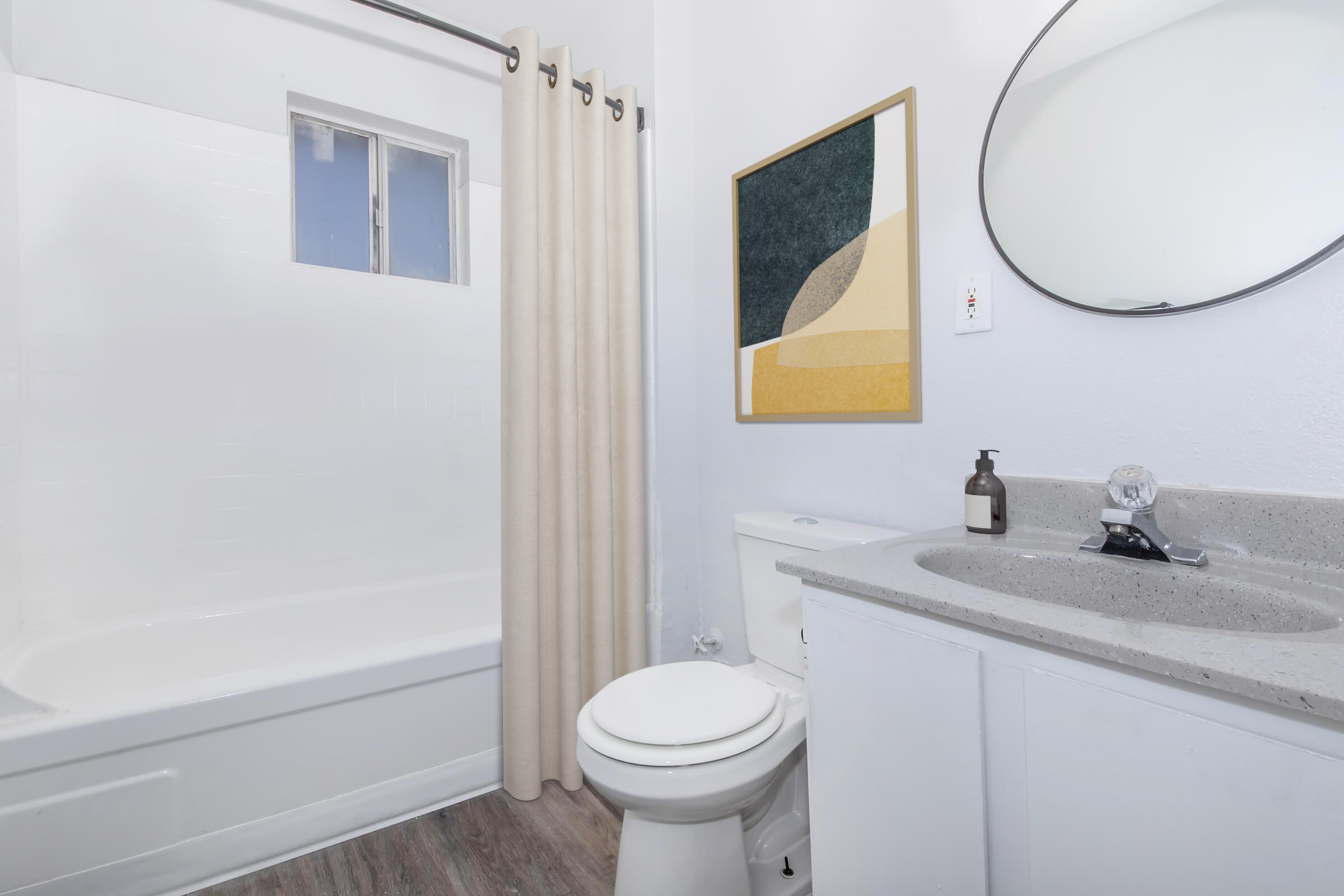 A clean and modern bathroom featuring a white bathtub with a shower curtain, a toilet, and a sink with a gray countertop. A round mirror is above the sink, and an abstract artwork is mounted on the wall, adding a touch of color to the space. The flooring is a light wood finish.