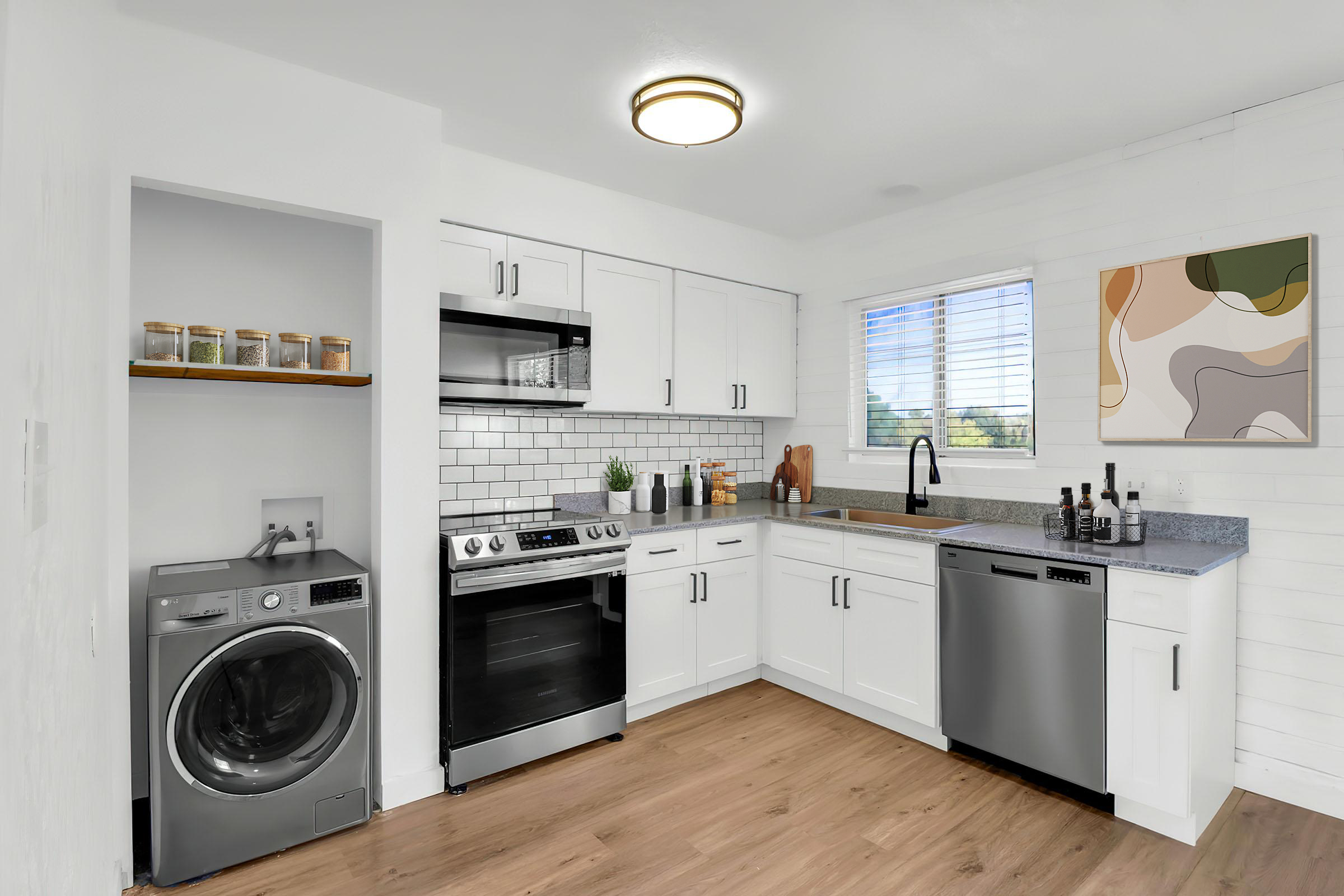 A modern kitchen featuring white cabinetry, stainless steel appliances, and a front-loading washing machine. The space includes a gray countertop, a tiled backsplash, and a window with natural light. Decorative items on the counter and a colorful abstract wall art piece add a stylish touch.
