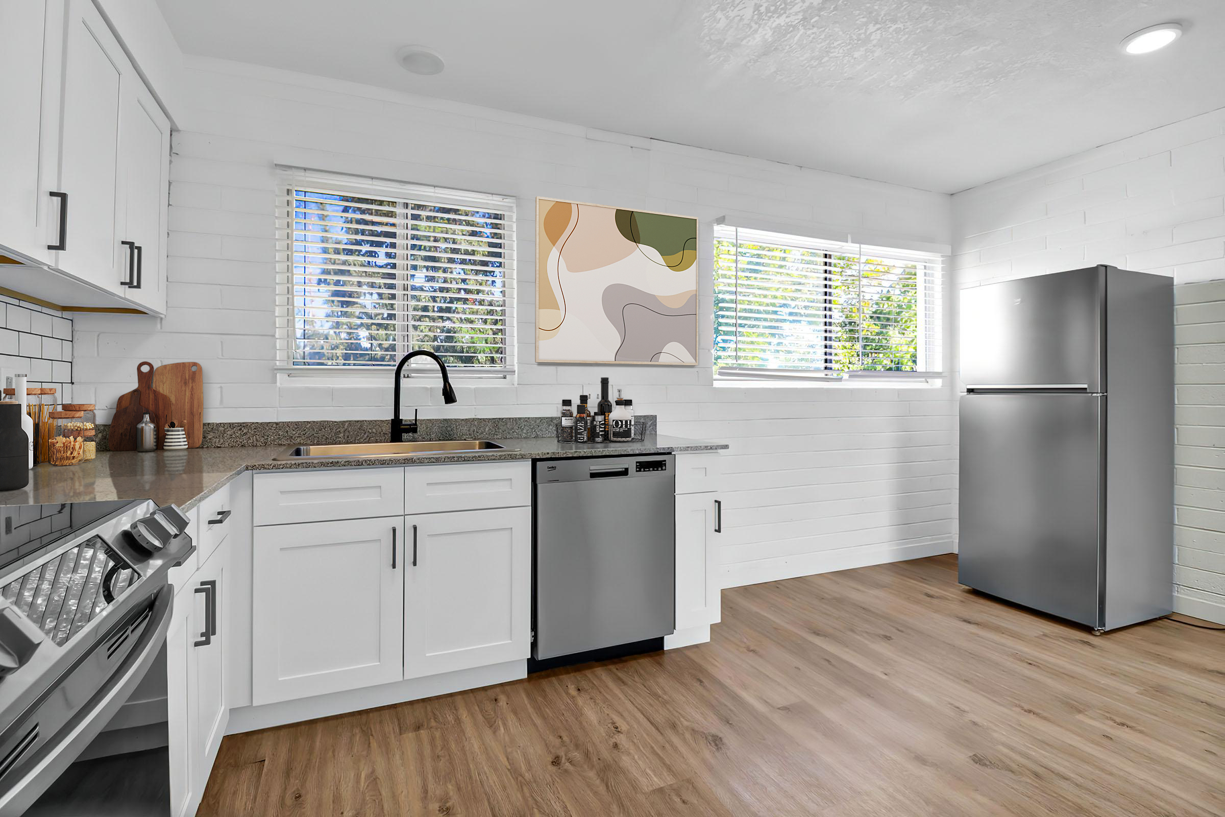 Modern kitchen featuring white cabinetry, a stainless steel refrigerator, and a gray dishwasher. Sunlight filters through two windows, illuminating a colorful abstract wall art piece. The countertop includes a sink, a cutting board, and various kitchen supplies, set against a backdrop of light wood flooring.