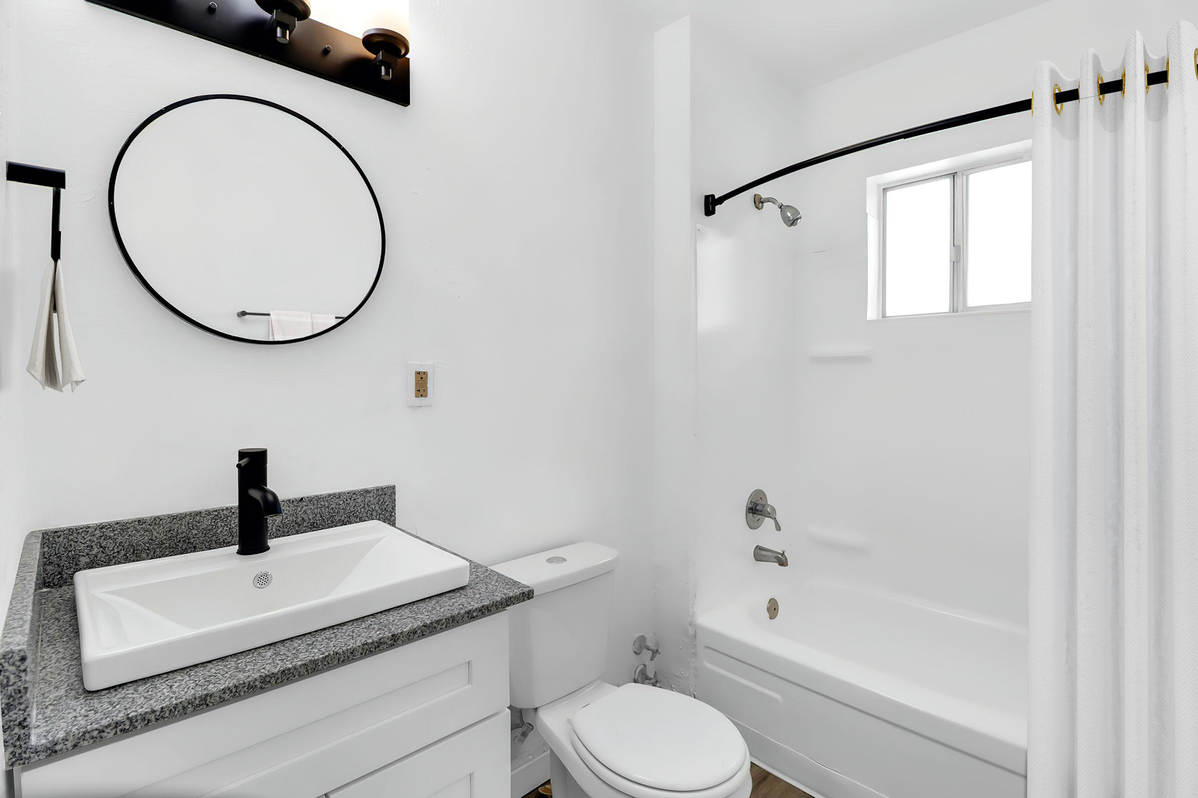 Modern bathroom featuring a sleek design with white walls, a round mirror above a stylish sink with a black faucet, a white toilet, and a bathtub with a shower curtain. Natural light enters through a window, enhancing the clean and minimalist aesthetic.
