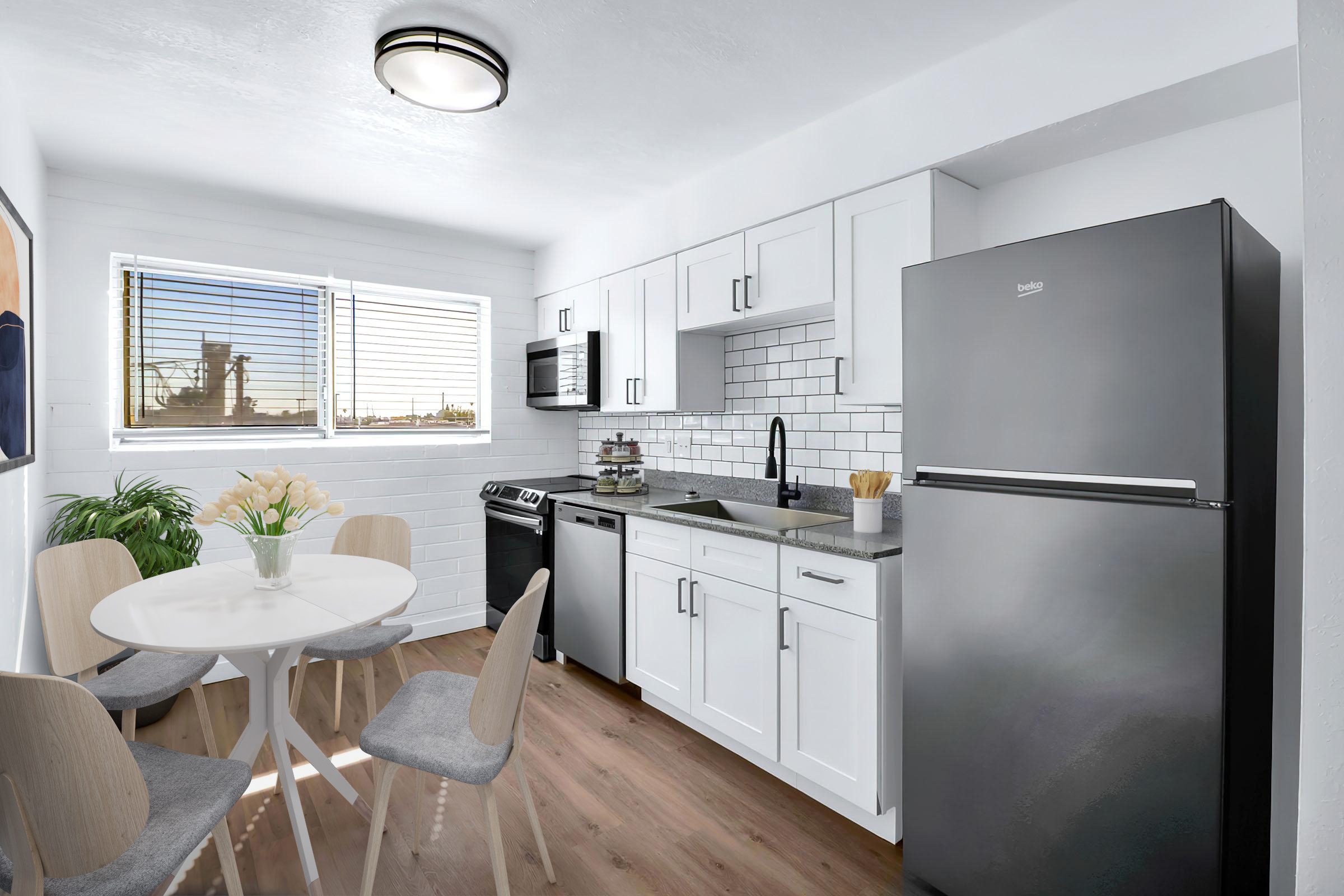 Modern kitchen featuring white cabinetry, stainless steel appliances, and a round dining table with light wood chairs. A window allows natural light to brighten the space, with a view of an outdoor industrial scene. The floor is a warm wood laminate, adding a cozy touch to the contemporary design.