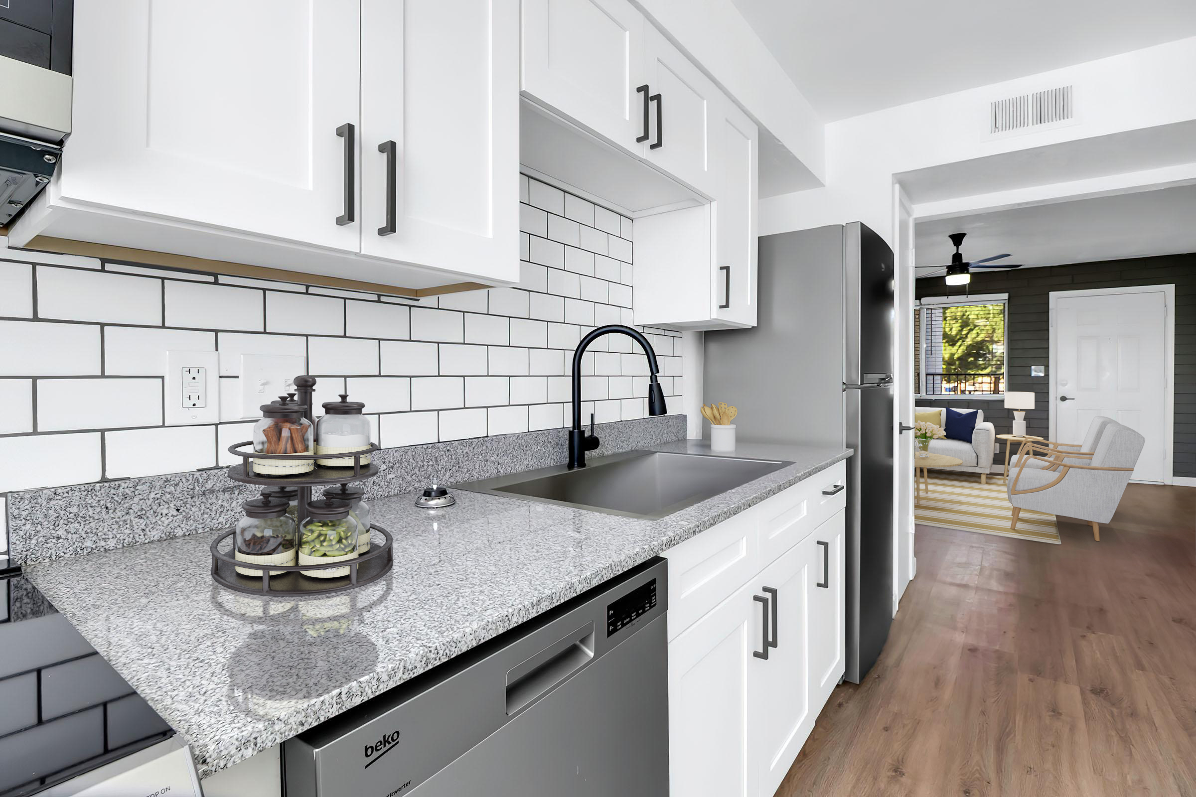 Modern kitchen with white cabinets, a grey granite countertop, and a stainless steel farmhouse sink. Features a decorative tiered stand with snacks, a dishwasher, and an open living area in the background, showcasing a comfortable chair and a ceiling fan. Bright and airy atmosphere with natural light.
