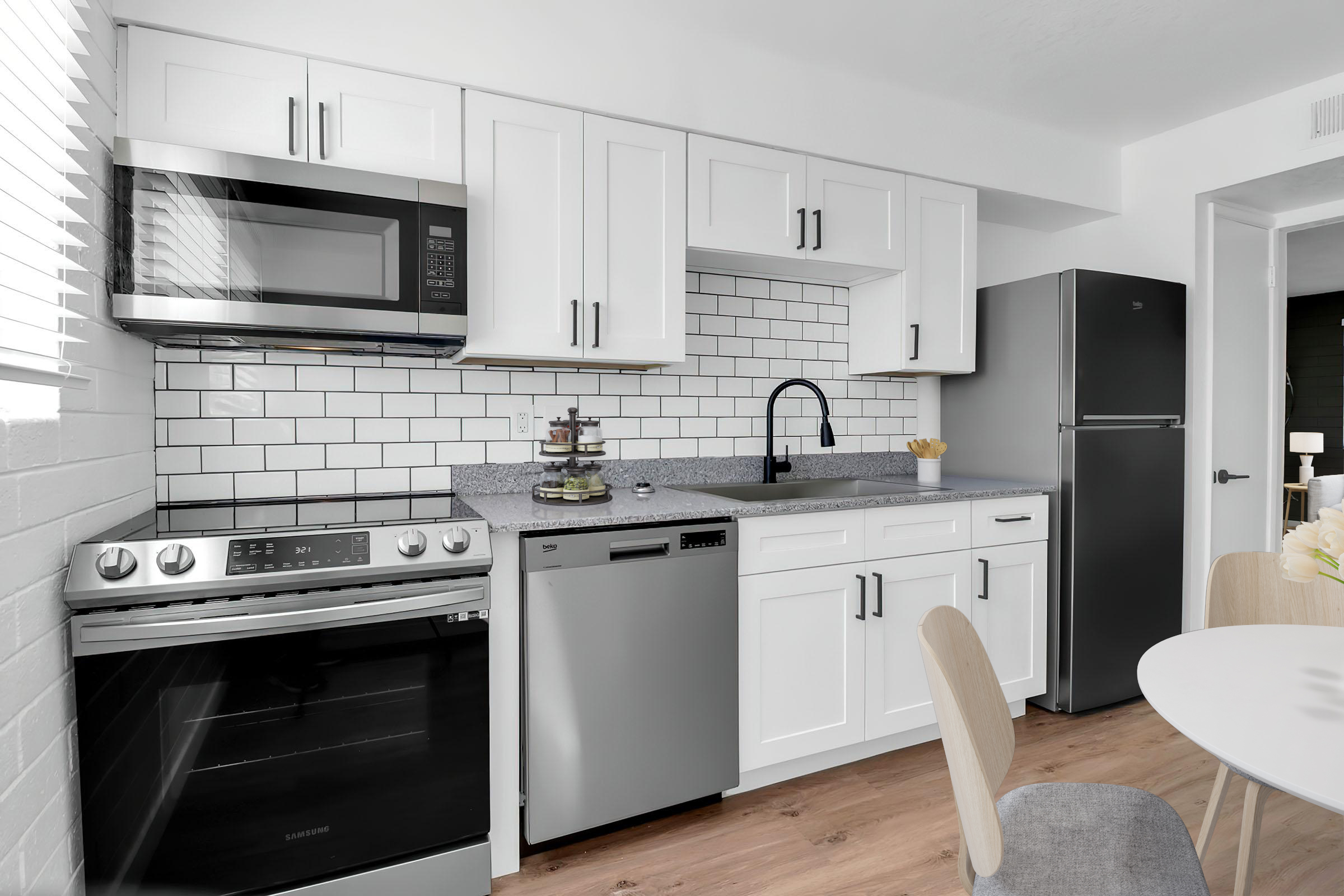 Modern kitchen featuring white cabinetry, stainless steel appliances including a microwave, oven, dishwasher, and refrigerator. The countertops are gray, with a stylish black faucet. Subway tile backsplash adds a sleek touch. A round dining table with light wooden chairs is visible, enhancing the clean, contemporary design.
