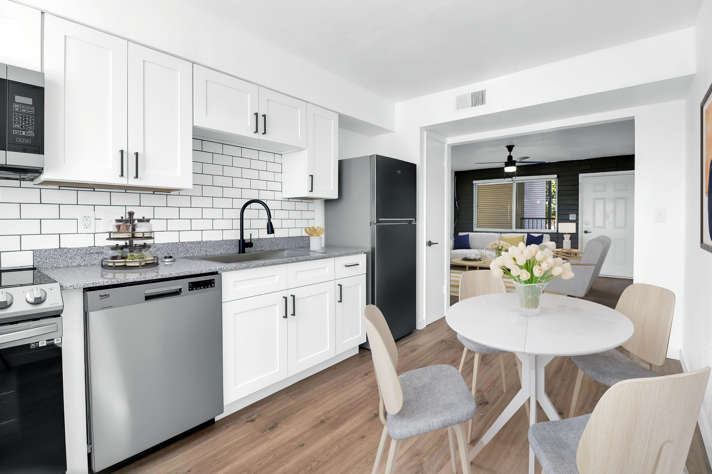 Modern kitchen featuring white cabinetry, a gray countertop, stainless steel appliances including a dishwasher, and a black faucet. A round dining table with light wooden chairs sits in the foreground, and a living area is visible in the background with large windows and a fan overhead.