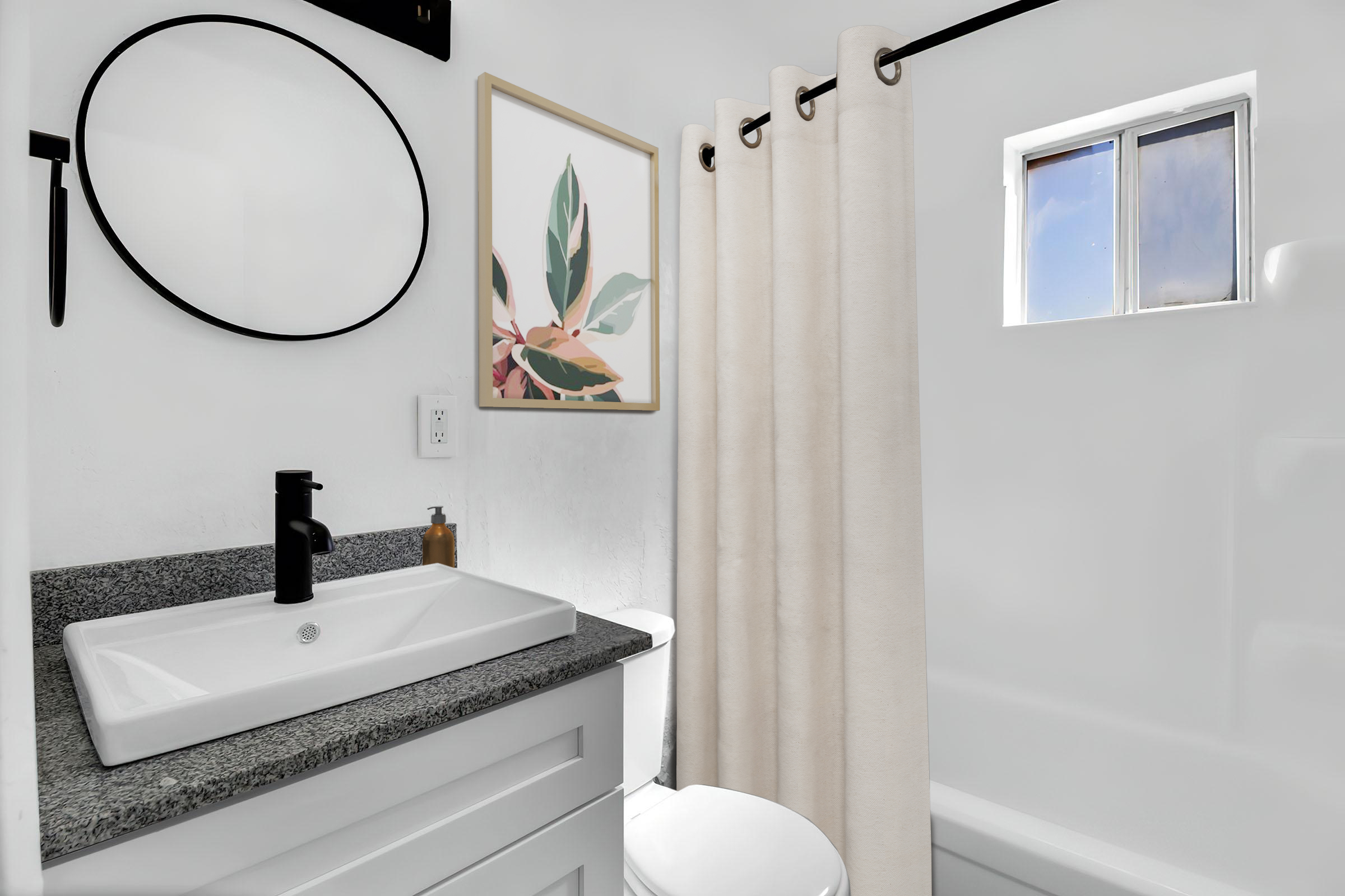 A modern bathroom featuring a circular mirror above a sink with a black faucet, a white shower curtain, a small window letting in natural light, and a decorative framed artwork of leaves on the wall. The space has clean lines and a minimalist design.