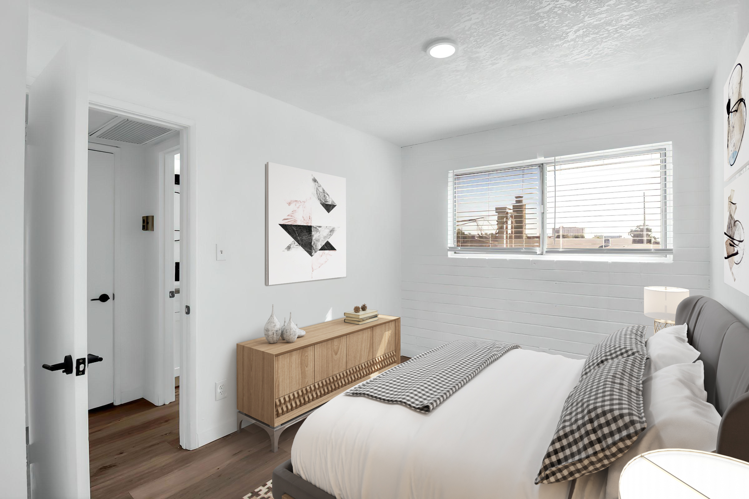 A modern bedroom featuring a gray upholstered bed with white linens, a wooden dresser with decorative items, and a large piece of abstract art on the wall. Natural light streams in through a window with white blinds, providing a bright and inviting atmosphere. A doorway leads to another room.