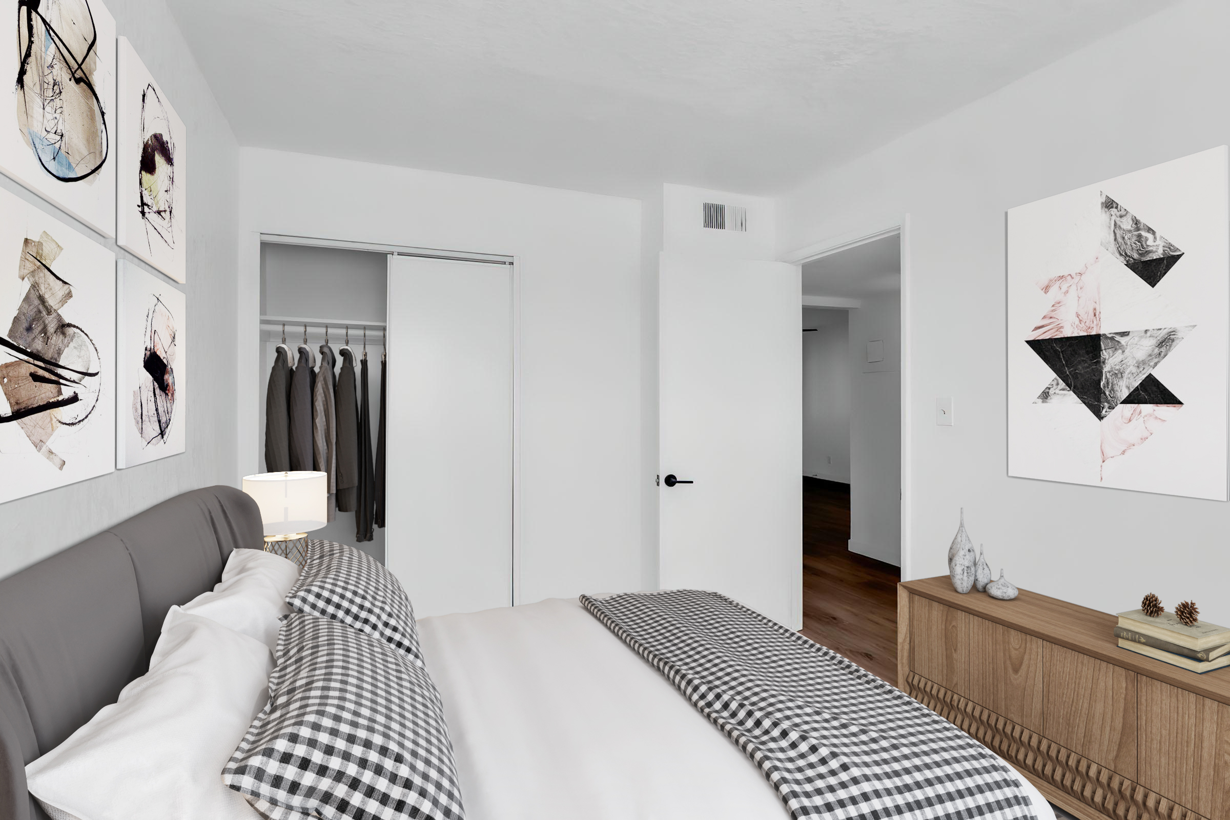 A modern bedroom featuring a bed with gray and white bedding, adorned with decorative pillows. The walls are painted white and decorated with abstract artwork. A wooden dresser is positioned beside the bed, and there is a closet with hanging clothes visible. Natural light illuminates the space from a doorway.