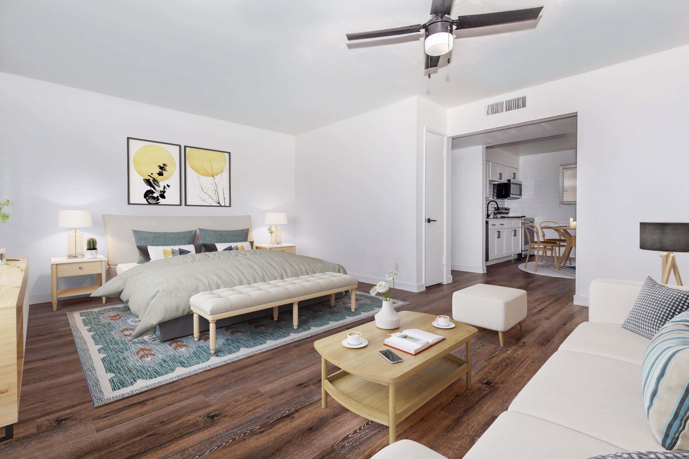 A modern, spacious bedroom featuring a large bed with a light gray comforter, a cozy seating area, and a stylish coffee table. Two framed prints of yellow leaves adorn the wall. Soft lighting from bedside lamps adds warmth, while an adjoining kitchen is visible in the background. The wooden floor enhances the inviting ambiance.