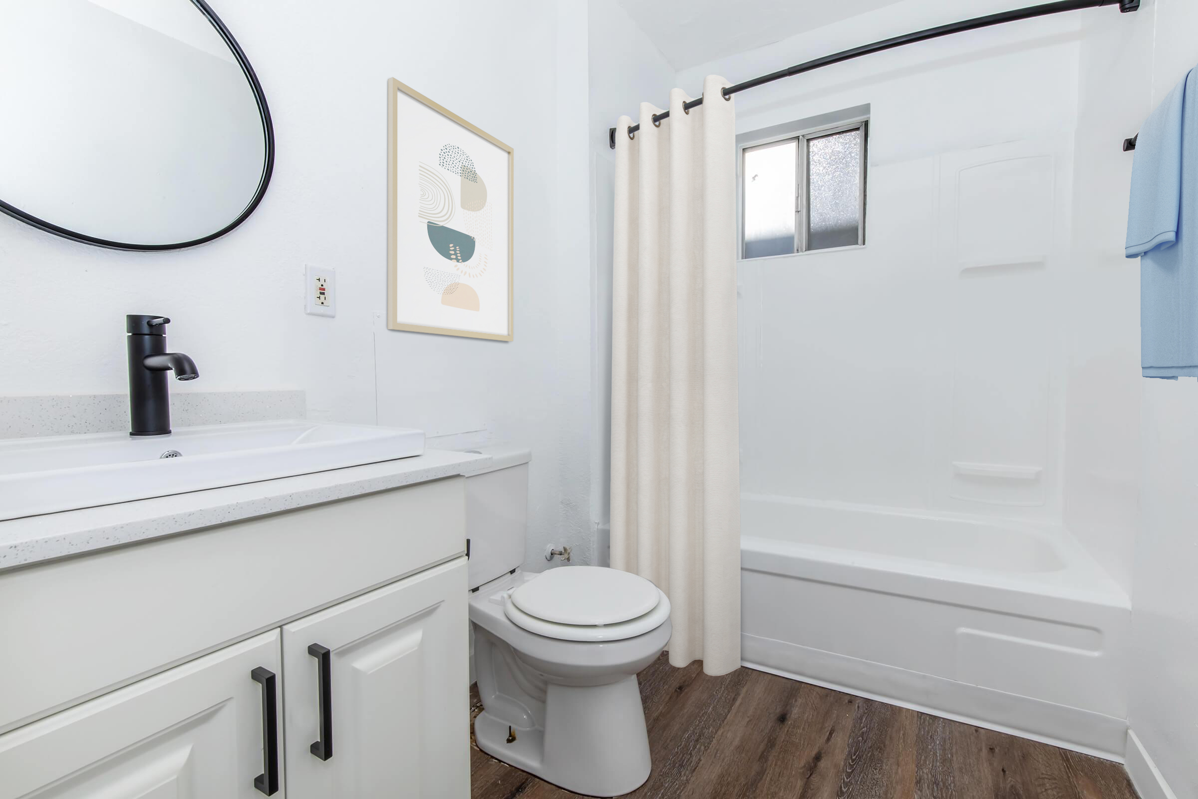 A modern bathroom featuring a white bathtub and shower, a black faucet, a round mirror, and a light-colored shower curtain. The walls are painted white, and there is a framed abstract art piece on the wall. A blue towel hangs on the side, with wood-like flooring completing the design.