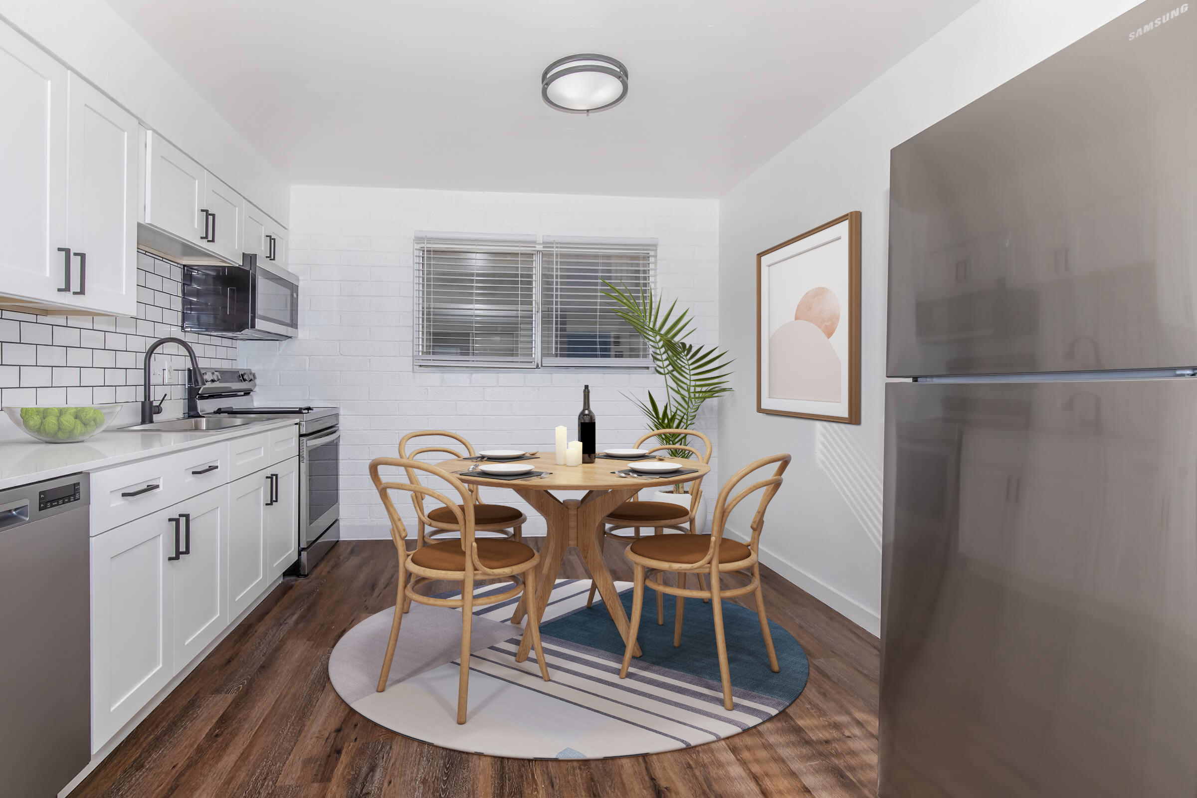 Modern kitchen with white cabinets, stainless steel appliances, and a round wooden dining table surrounded by chairs. A bottle of wine and plates are set on the table. The space features a plant for decoration and a contemporary art piece on the wall, with large windows allowing natural light.