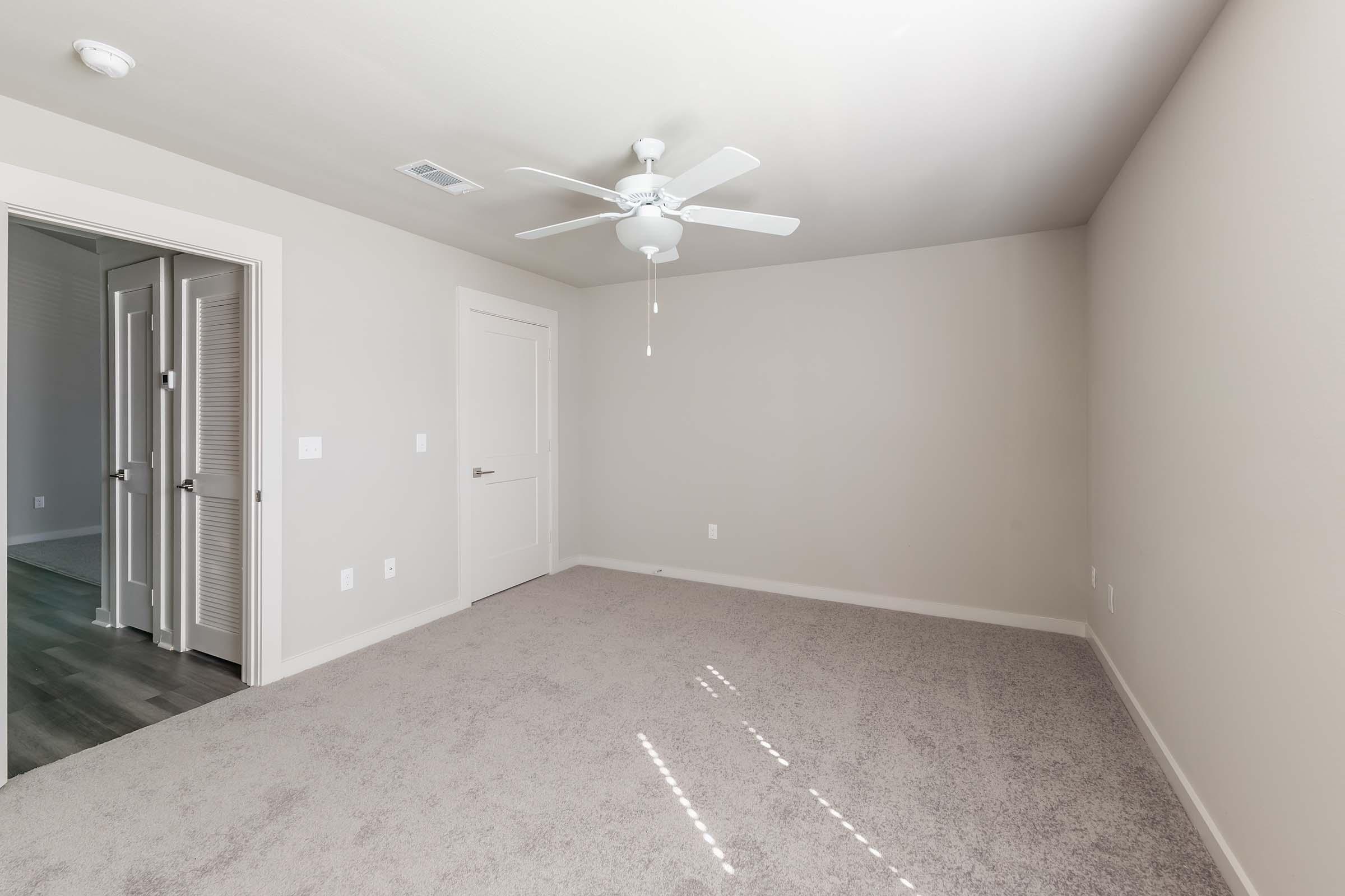 A spacious, empty room with light beige walls and a ceiling fan. It features carpeted flooring and a door leading to another area. There are two closed doors visible, likely leading to storage or a closet. Soft natural light filters in through a window, illuminating the clean, minimalist space.