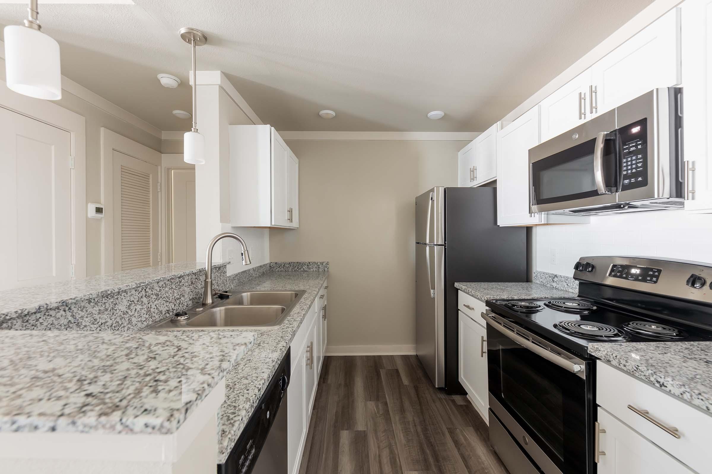 Modern kitchen featuring white cabinetry, granite countertops, stainless steel appliances including a refrigerator, microwave, and oven. The space has a neutral color palette, with pendant lights above the counter and a spacious layout that emphasizes functionality.