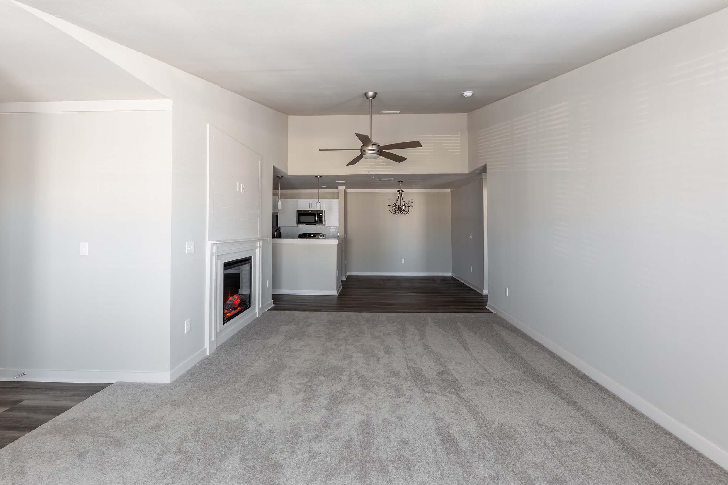 A spacious, empty living room featuring light gray carpet, a ceiling fan, and a modern fireplace. In the background, there is an opening leading to a kitchen area with a microwave. The walls are painted light gray, and the room is well-lit with natural light.