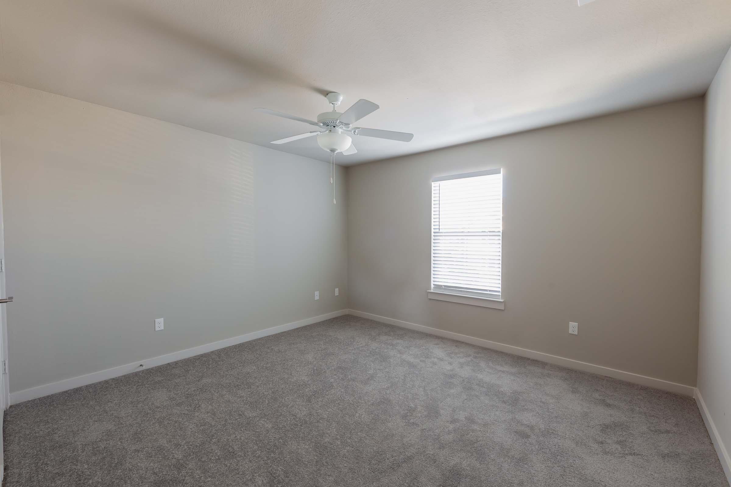 A vacant room featuring light gray walls and carpet. It includes a ceiling fan and a single window with blinds providing natural light. The space appears clean and simple, suitable for various uses.