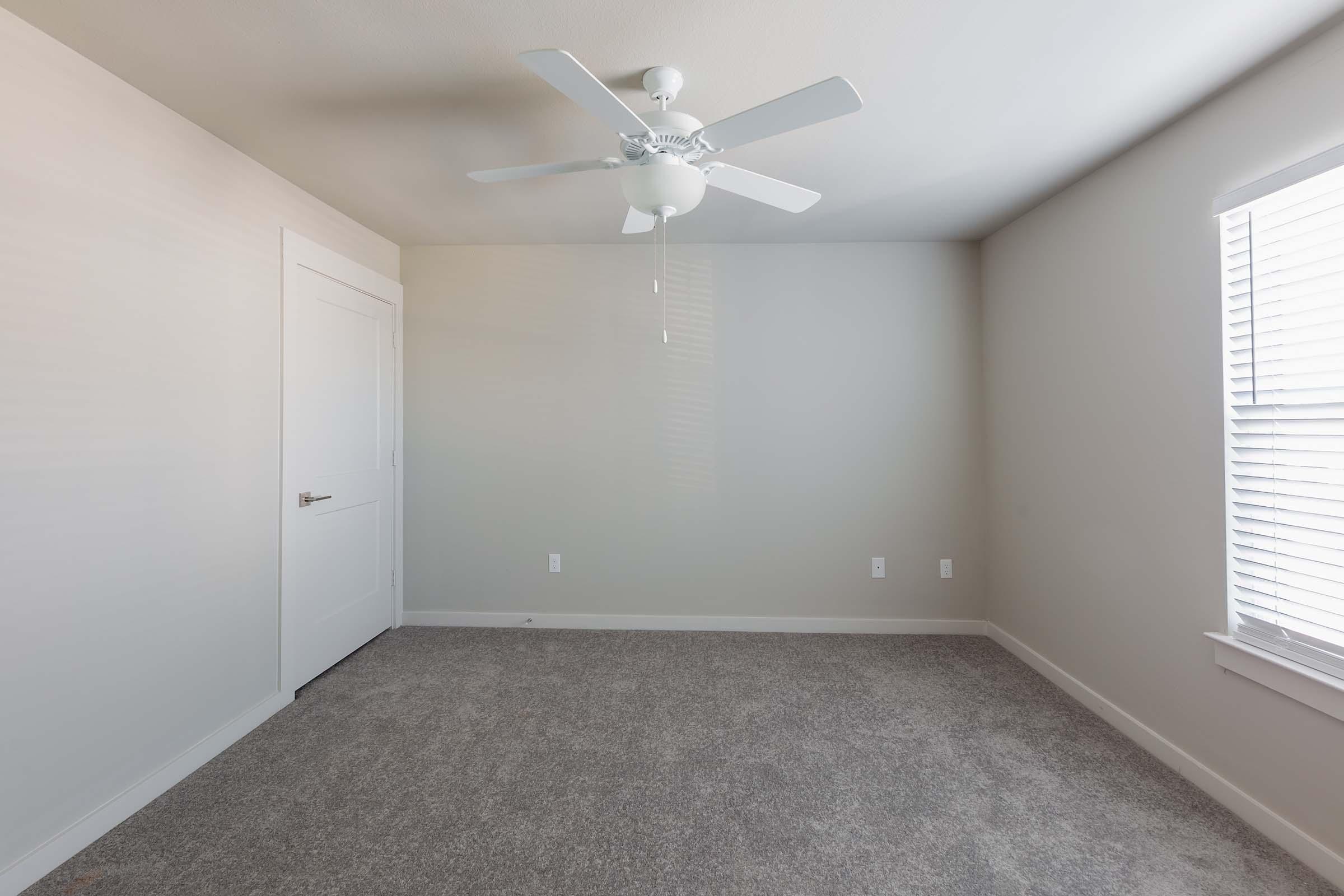 A spacious, empty room with light gray walls and newly installed carpet. It features a white ceiling fan, a white door, and a window with blinds that allows natural light to enter. The room is uncluttered, making it ideal for various uses or personal decoration.