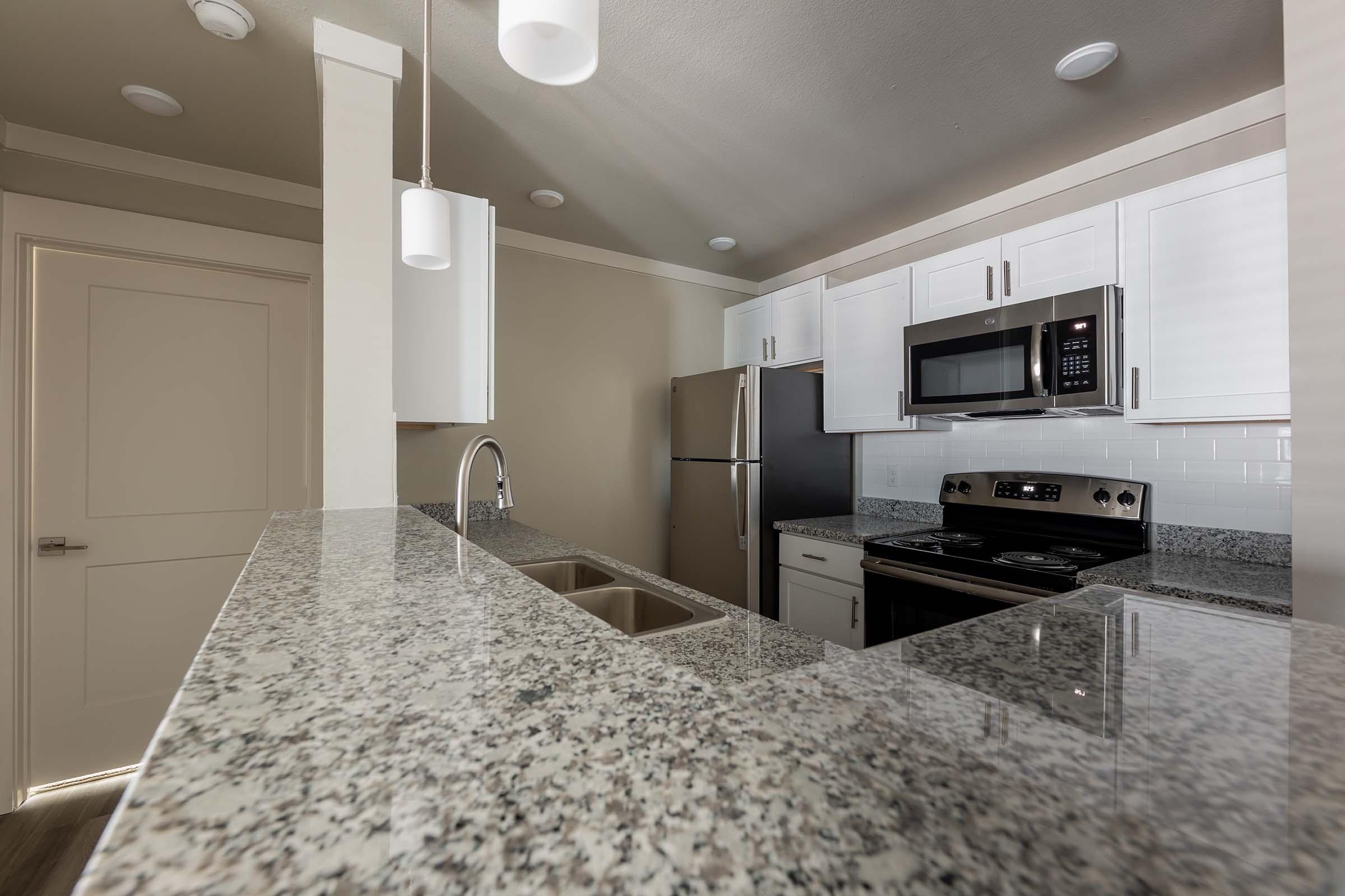 Modern kitchen featuring a granite countertop with a double sink, stainless steel appliances including a refrigerator and microwave, and white cabinetry. The room is well-lit with pendant lights and recessed ceiling lights, creating a bright and inviting ambiance.