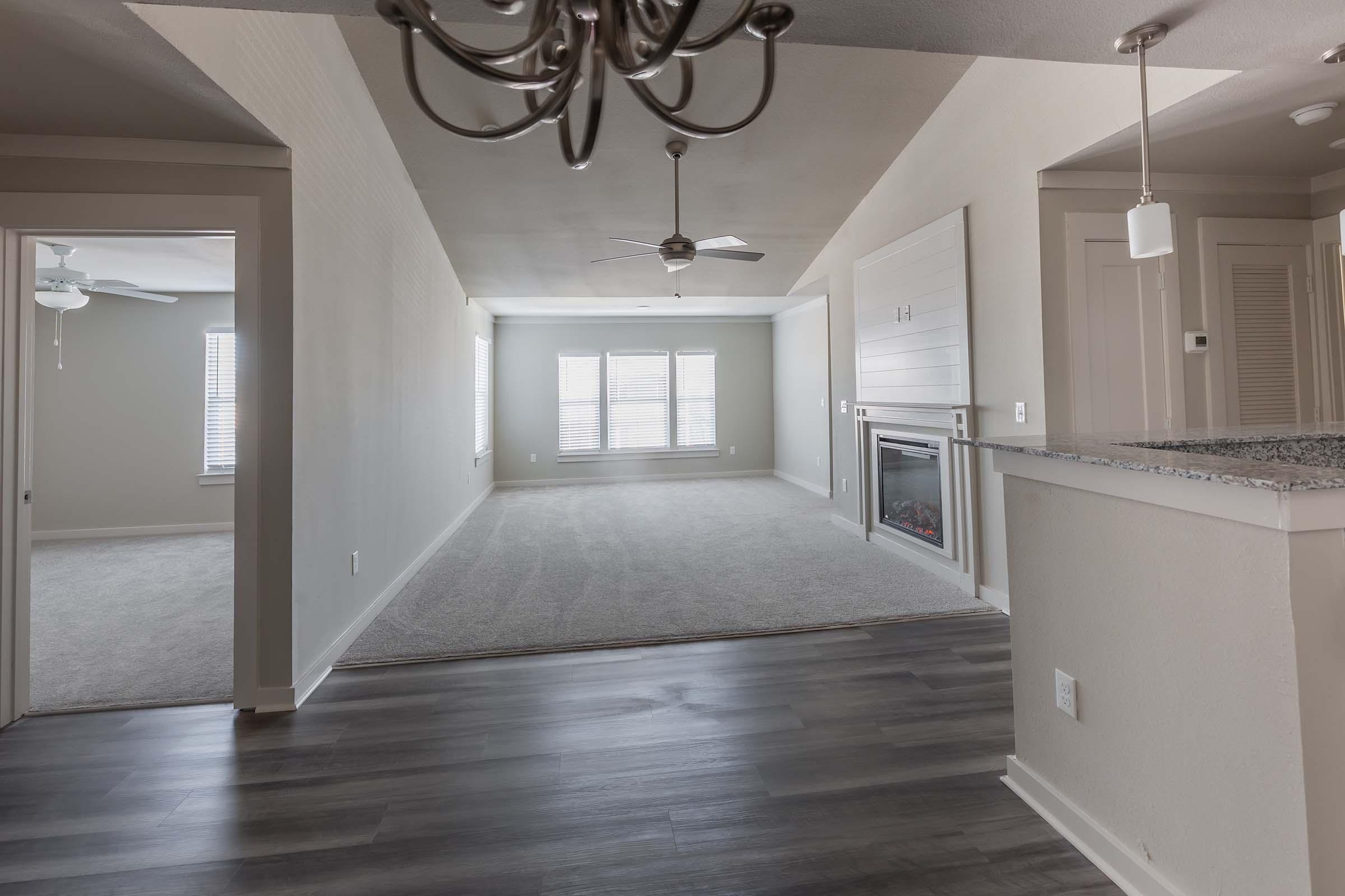 A spacious living area featuring a ceiling fan, large windows with blinds, and a decorative electric fireplace. The floor is covered in light carpet, and there is a view into an adjacent room. The walls are painted in a light color, and the flooring transitions to dark hardwood.