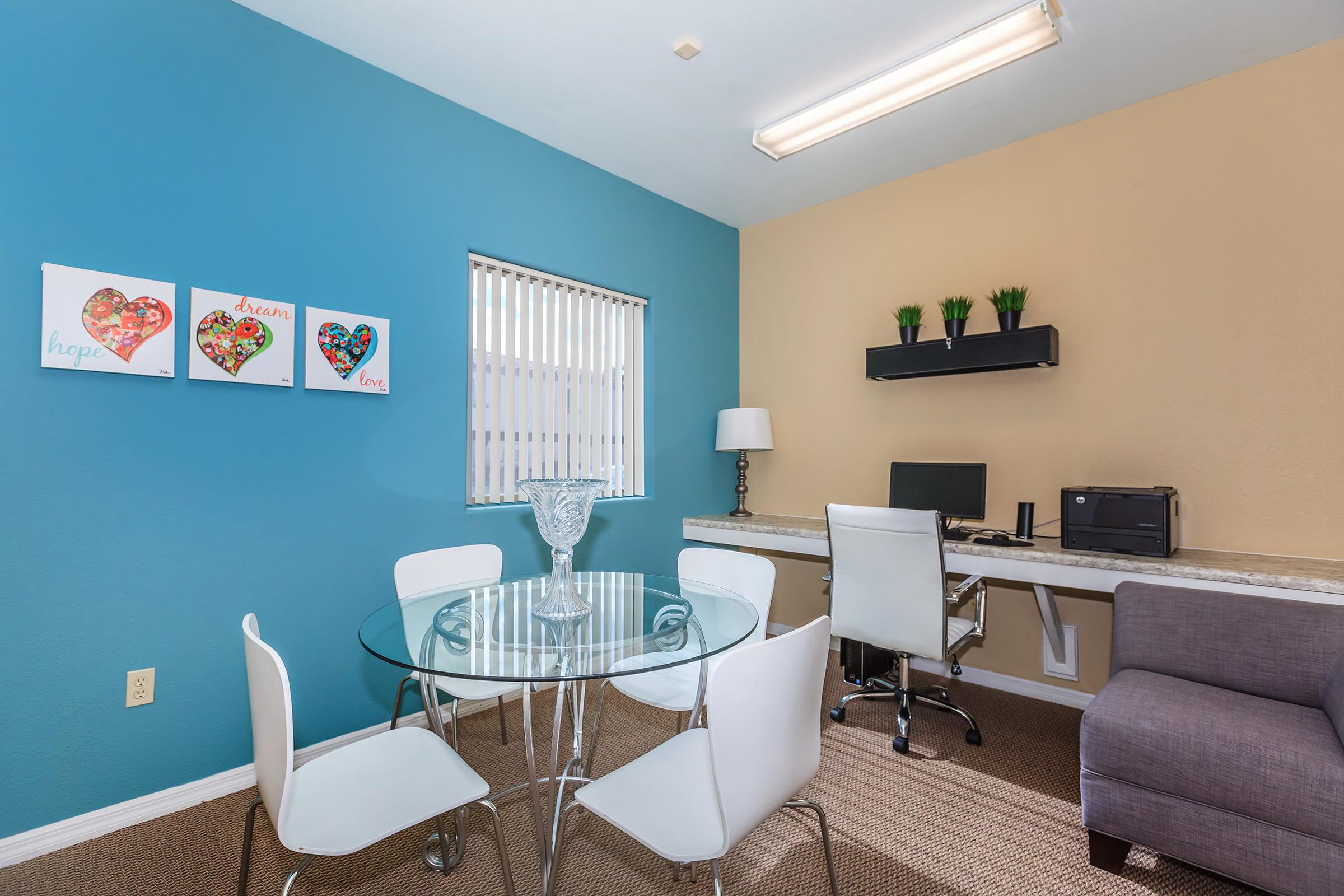 A small office space featuring a glass dining table with four white chairs, a wall with colorful heart artwork, a desk with a computer and a lamp, a microwave, and a gray chair.