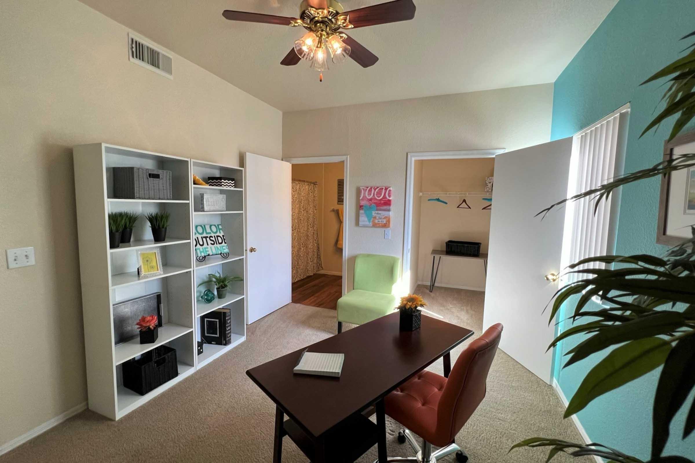 A bright and inviting office space featuring a desk with a notebook, two chairs, a white bookshelf with decorative items, and an open door leading to a closet.