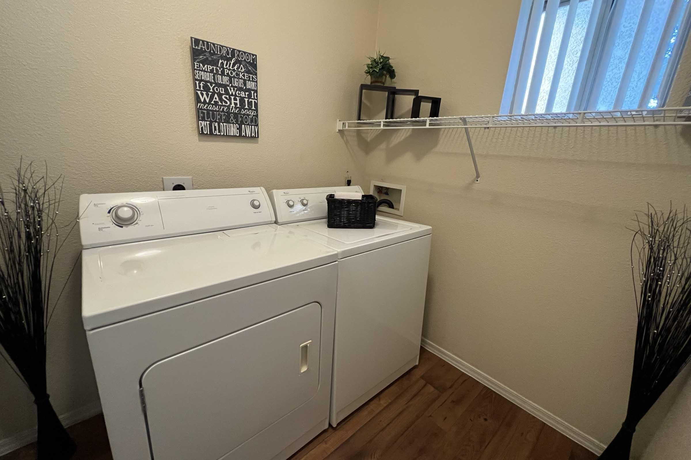 A clean laundry room featuring a white washer and dryer, a wall-mounted shelf with a decorative plant, and a framed wall sign with laundry instructions.